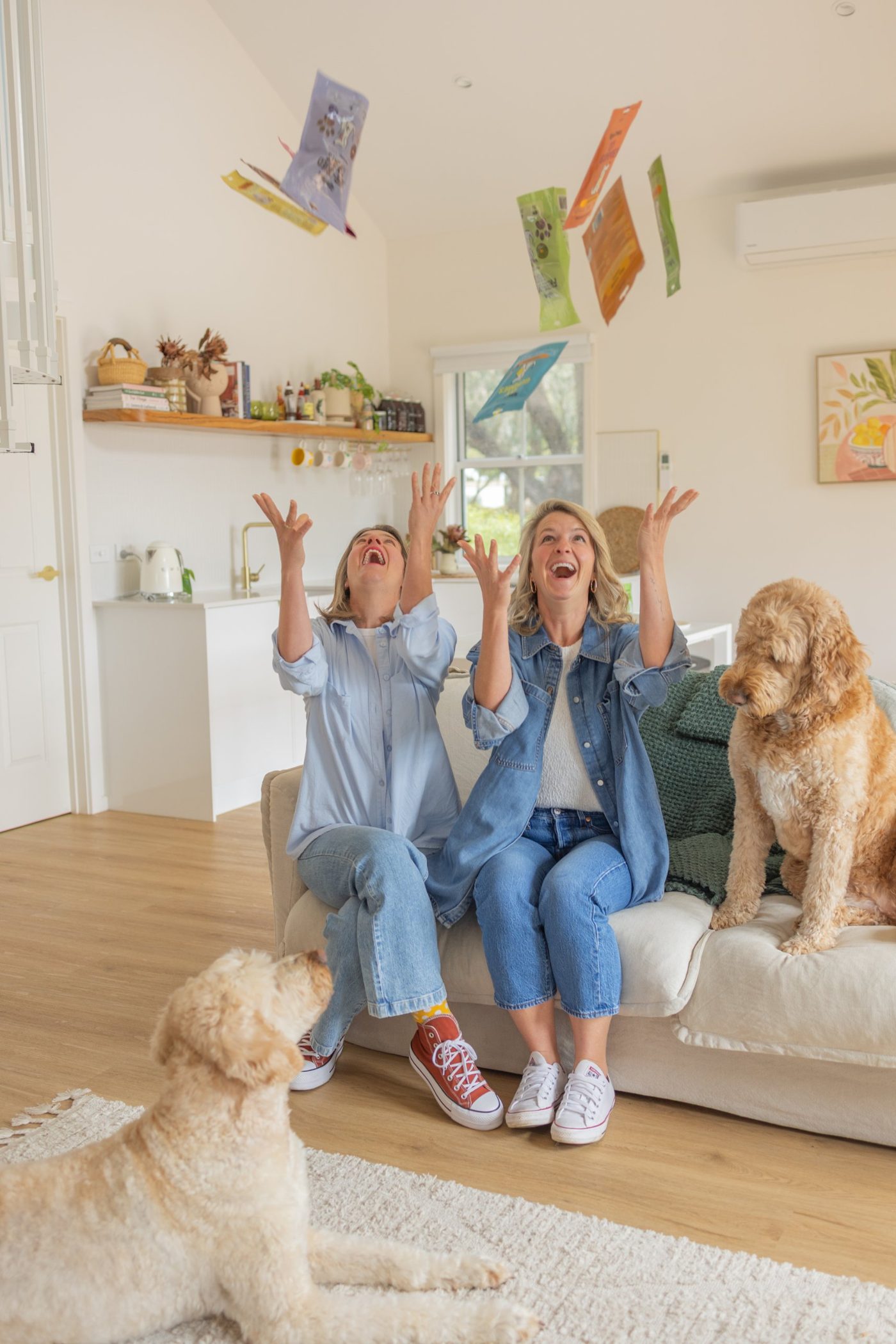 Alana and a friend laughing on a couch with two dogs, tossing product packets in the air