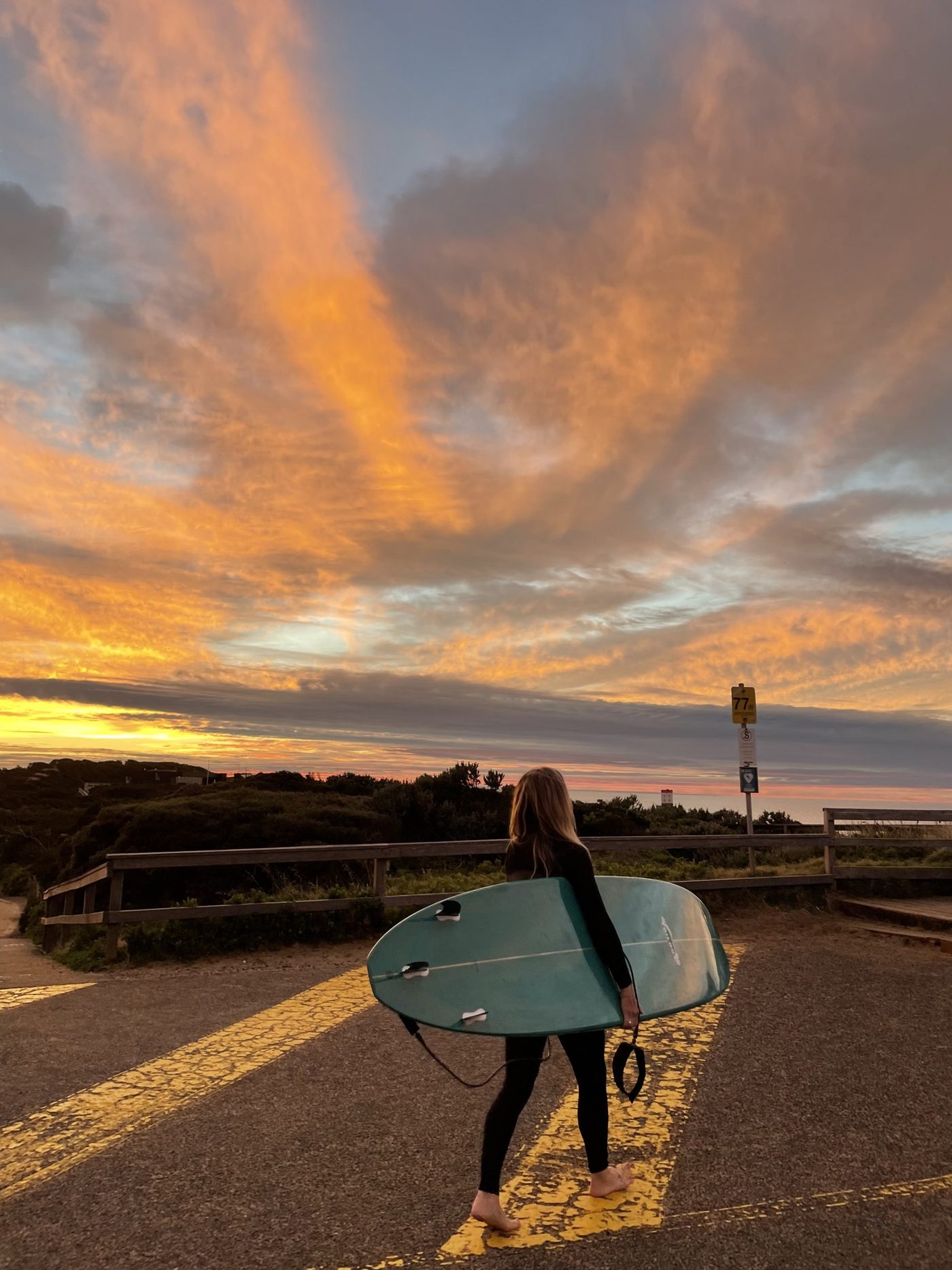 Alana carrying a surfboard across a car park under a sunrise sky